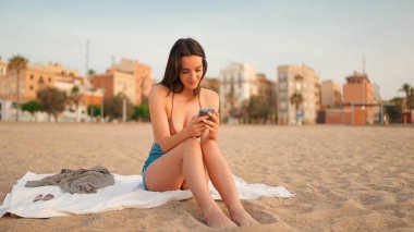 İnce, kahverengi saçlı, uzun saçlı bir kadın kumsalda cep telefonu kullanıyor. Güzel, gülümseyen kız akıllı telefona bilgi kaydırıyor.. 