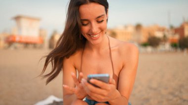 İnce, kahverengi saçlı, uzun saçlı bir kadın kumsalda cep telefonu kullanıyor. Güzel, gülümseyen kız akıllı telefona bilgi kaydırıyor.. 