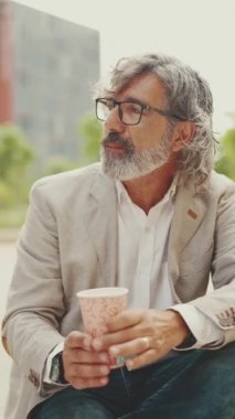VERTICAL VIDEO: Mature businessman with beard in eyeglasses wearing gray jacket holding takeaway coffee. Middle aged manager enjoying morning coffee while sitting outside the office