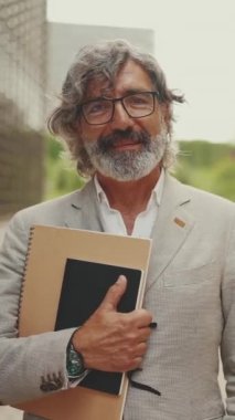 Gözlüklü, sakallı, gri ceketli, gülümseyen olgun iş adamı etrafına bakıyor ve modern binaların önünden geçiyor.