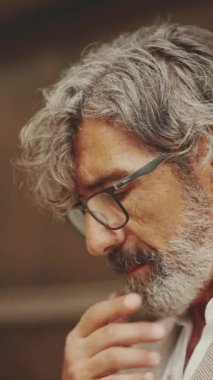 VERTICAL VIDEO: Close-up profile of mature businessman with beard in eyeglasses wearing gray jacket rubs his chin in thought