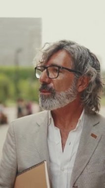 Gözlüklü, gri ceketli, iş belgeleriyle dolu bir klasörü olan olgun bir işadamı, modern binaların önünden geçiyor.