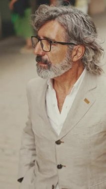 Gözlüklü, sakallı, gri ceketli, olgun bir iş adamı modern binaların önünden geçiyor.