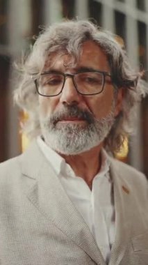 Gözlüklü, sakallı, gri ceketli, olgun bir iş adamı modern binaların önünden geçiyor. Orta yaşlı yönetici kol saatine bakıyor.
