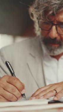 VERTICAL VIDEO: Mature businessman with beard in eyeglasses, user on laptop computer and taking notes, sitting in outdoor cafe.Man sitting at table in cafe in nature Mobile office freelancer