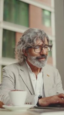 Gözlüklü, gri ceketli, sakallı, olgun bir işadamı kafede oturup kahve içiyor. Bilgisayarda çalışan başarılı bir adam dışarıda, kafede oturuyor.