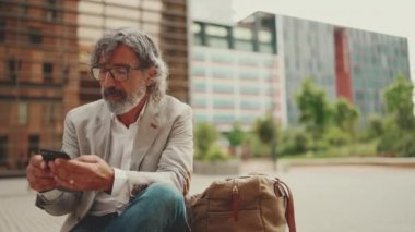 Gözlüklü, gri ceketli, sakallı, olgun bir işadamı cep telefonu kullanıyor. Orta yaşlı yönetici ofisin dışında otururken akıllı telefonuyla bilgi kaydırıyor.