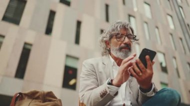 Mature businessman with beard in eyeglasses wearing gray jacket holds cellphone in his hand and looks around pondering