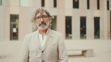 Gözlüklü, gri ceketli, sakallı, olgun bir işadamı kollarını kavuşturup kameraya bakıyor. Ofisin dışında dikilirken orta yaşlı yönetici kameraya poz veriyor.