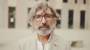 Gözlüklü, gri ceketli, sakallı yetişkin iş adamının yakın çekimi kameraya bakıyor. Ofisin dışında dikilirken orta yaşlı yönetici kameraya poz veriyor.