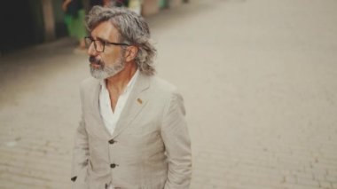Gözlüklü, gri ceketli, olgun bir iş adamı modern binaların önünden geçiyor.