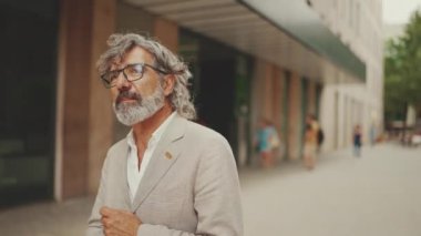 Gözlüklü, gri ceketli, sakallı, olgun bir işadamı etrafına bakıyor ve modern binaların önünden geçiyor.