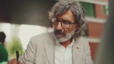 Sakallı, olgun iş adamı, dizüstü bilgisayarı kullanan ve notlar alan açık bir kafede oturan. Başarılı bir adam doğadaki kafede oturuyor. Mobil ofis çalışanı.