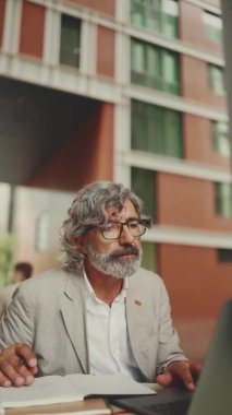 VERTICAL VIDEO: Mature businessman with beard in eyeglasses, user on laptop computer and taking notes, sitting in outdoor cafe.Man sitting at table in cafe in nature Mobile office freelancer