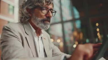 Clouse up, mature businessman with beard in eyeglasses wearing gray jacket, working on tablet pc. Successful man sitting at table Mobile office freelancer