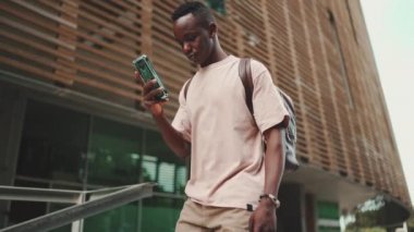 Young african student walks down the stairs outside of the university, using cellphone