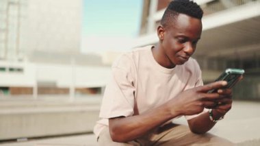 Genç Afrikalı öğrenci üniversitenin dışında oturuyor, cep telefonu kullanıyor, akıllı telefon ekranını dinliyor.