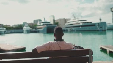 Young african guy relaxes sitting on bench in the port on yachts and ships background. Back view