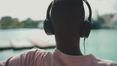 Clouse yukarı, genç Afrikalı adam limanda bankta oturuyor, kulaklıkla müzik dinliyor, şarkı söylüyor. Arka plan