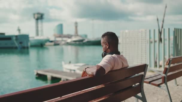 Young African Guy Relaxes Sitting Bench Port Yachts Ships Background — Stockvideo