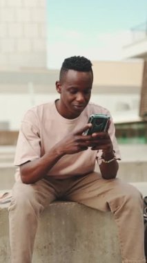 Genç Afrikalı öğrenci üniversitenin dışında oturuyor, cep telefonu kullanıyor, akıllı telefon ekranını dinliyor.