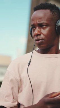 VERTICAL VIDEO: Young african student sitting outside of university wearing headphones, using phone, listening to music