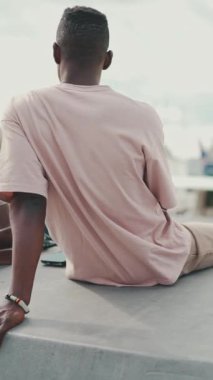 VERTICAL VIDEO: Young African guy is sitting on the embankment, against the background of yachts and ships. Back view