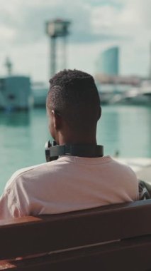 VERTICAL VIDEO: Young african guy relaxes sitting on bench in the port on yachts and ships background. Back view