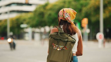 Uzun kahverengi saçlı, beyaz saçlı, omuzlarında sırt çantası olan sarı bandanalı hoş bir kadının profili yaya geçidi boyunca yürüyor.