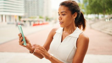 Beyaz bluz giyen güzel, bronzlaşmış kadın cep telefonu kullanıyor. Kız cep telefonuyla şehrin arka planında selfie video konferansı düzenliyor.