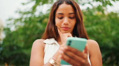 Uzun siyah saçlı, beyaz bluzlu güzel kız bankta oturuyor ve cep telefonu kullanıyor. Sosyal ağlarda cep telefonuyla yazan gülümseyen genç bir kadın..