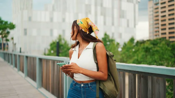 Cute tanned woman with long brown hair wearing white top and yellow bandana walks on bridge with backpack on her shoulder and cell phone in her hand. Girl using phone