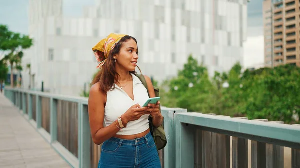 Cute tanned woman with long brown hair wearing white top and yellow bandana walks on bridge with backpack on her shoulder and cell phone in her hand. Girl using phone