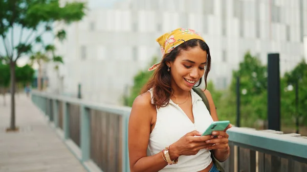 Cute tanned woman with long brown hair wearing white top and yellow bandana walks on bridge with backpack on her shoulder and cell phone in her hand. Girl using phone