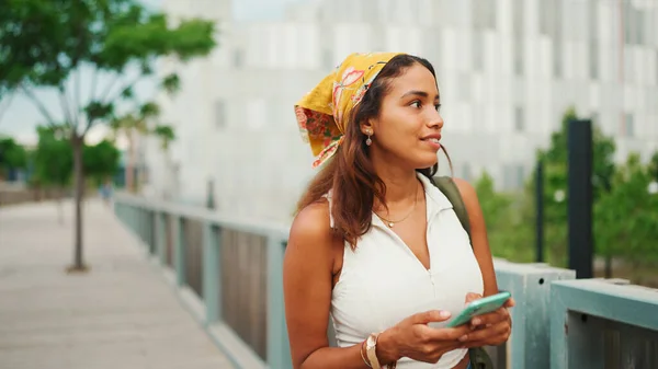 Cute tanned woman with long brown hair wearing white top and yellow bandana walks on bridge with backpack on her shoulder and cell phone in her hand. Girl using phone