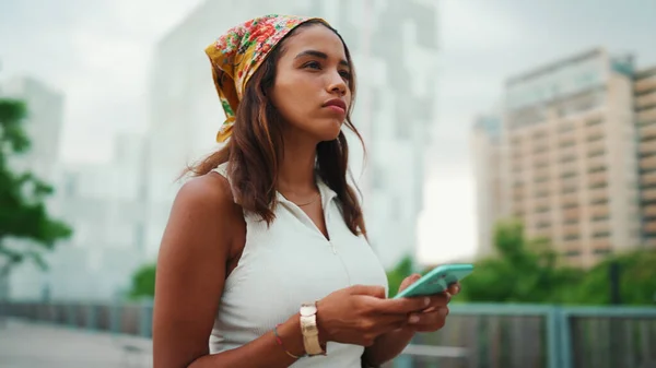 Cute tanned woman with long brown hair wearing white top and yellow bandana walks on bridge with backpack on her shoulder and cell phone in her hand. Girl using phone