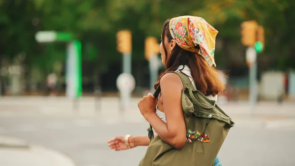 Profile of cute tanned woman with long brown hair in white top and yellow bandana with backpack on her shoulders is walking along pedestrian crossing