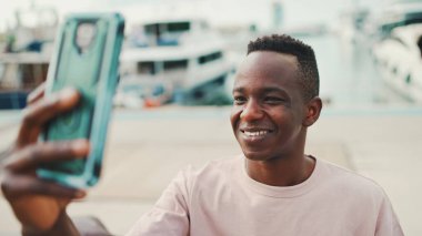Genç Afrikalı adam, arka planda yatlar ve gemilerle barajda duran telefonu kullanıyor ve video görüşmesi yapıyor.
