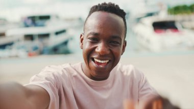 Genç Afrikalı adam, arka planda yatlar ve gemilerle barajda duran telefonu kullanıyor ve video görüşmesi yapıyor.