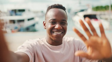 Genç Afrikalı adam, arka planda yatlar ve gemilerle barajda duran telefonu kullanıyor ve video görüşmesi yapıyor.