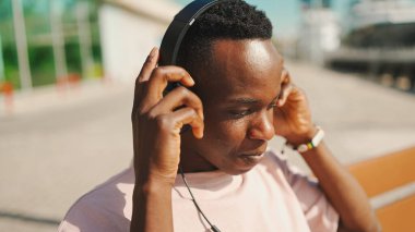 Gülümseyen Afrikalı genç adam limanda bankta oturuyor, kulaklıkla müzik dinliyor, birlikte şarkı söylüyor.