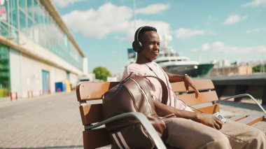 Gülümseyen Afrikalı genç adam limanda bankta oturuyor, kulaklıkla müzik dinliyor, birlikte şarkı söylüyor.