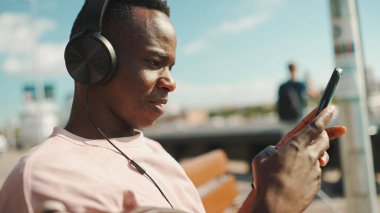 Genç Afrikalı adam bankta oturup, kulaklıkla müzik dinliyor, cep telefonu kullanıyor, şarkı söylüyor.