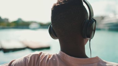 Clouse yukarı, genç Afrikalı adam ferahlıyor limanda yat ve gemi arka planında bankta oturuyor. Arka plan