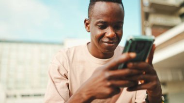 Genç Afrikalı öğrenci üniversitenin dışında oturuyor, cep telefonu kullanıyor, akıllı telefon ekranını dinliyor.