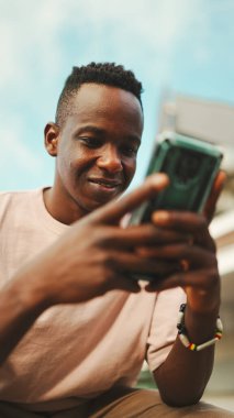 Genç Afrikalı öğrenci üniversitenin dışında oturuyor, cep telefonu kullanıyor, akıllı telefon ekranını dinliyor.