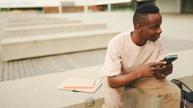Genç Afrikalı öğrenci üniversitenin dışında oturuyor, cep telefonu kullanıyor, akıllı telefon ekranını dinliyor.