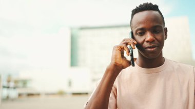 Üniversitenin dışında oturan gülümseyen Afrikalı genç bir öğrenci cep telefonuyla konuşuyor.. 