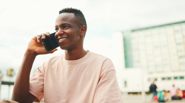 Üniversitenin dışında oturan gülümseyen Afrikalı genç bir öğrenci cep telefonuyla konuşuyor.. 