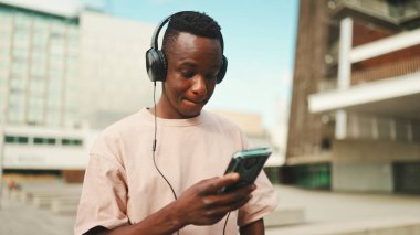 Üniversitenin dışında oturan genç Afrikalı öğrenci kulaklık takıyor, telefon kullanıyor, müzik dinliyor.